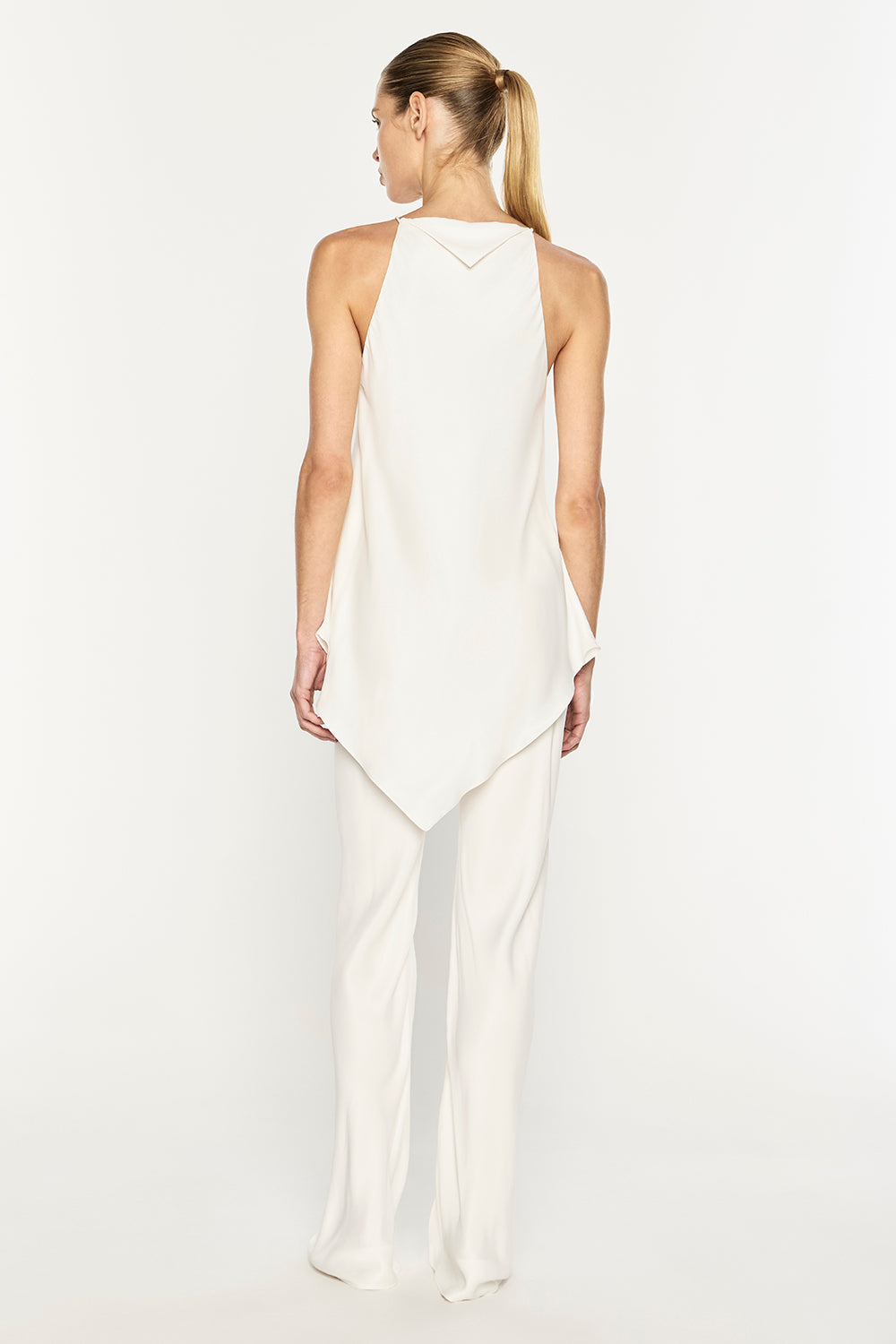 A woman viewed from the back wearing a sleeveless white halter top with an asymmetrical draped hem and matching wide-leg white pants, photographed against a clean white studio background.