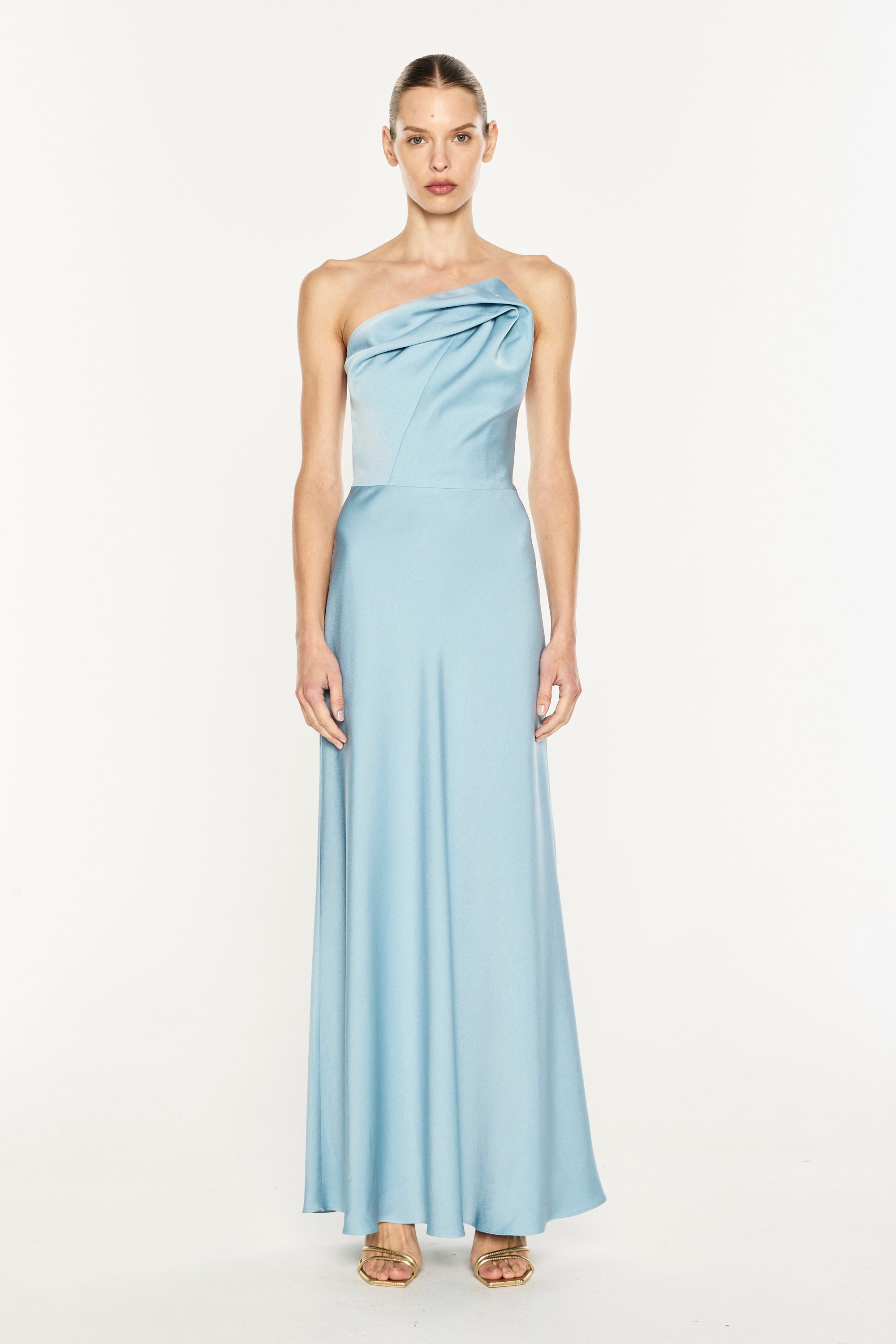 A model wearing a light blue one-shoulder satin maxi dress with a draped bodice and flowing skirt, standing against a plain white studio background.