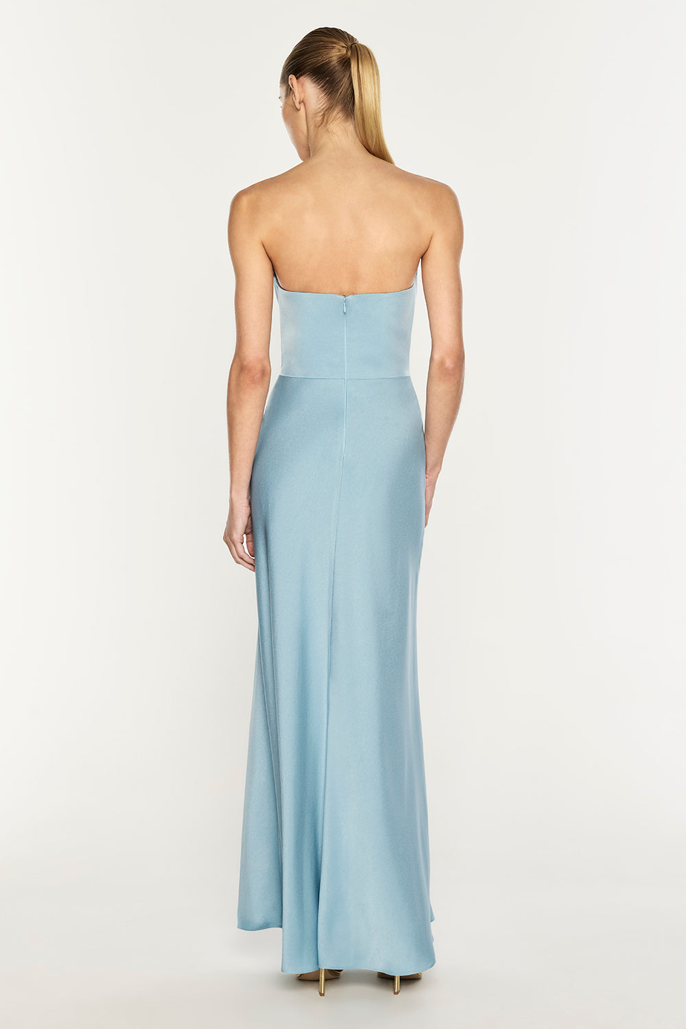 A woman viewed from the back wearing a strapless light blue floor-length gown against a plain white studio background.