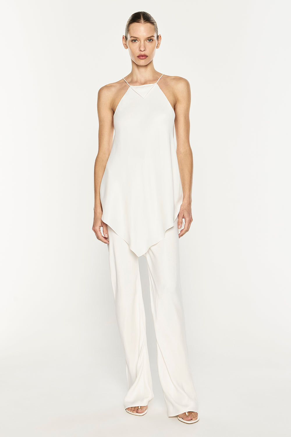 A model wearing a white sleeveless halter top with an asymmetrical draped hem and matching wide-leg trousers, photographed against a seamless white studio background.