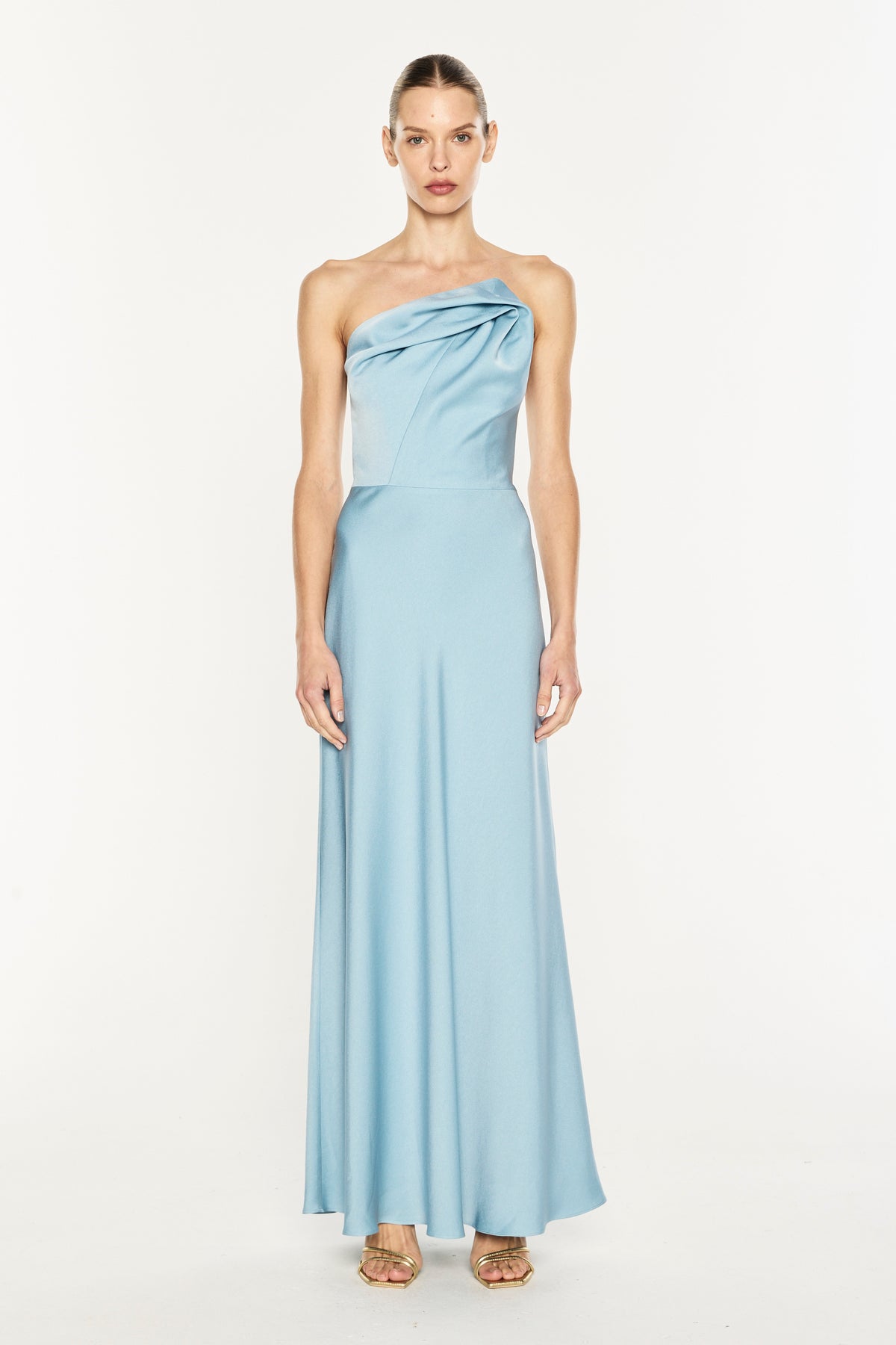A model wearing a light blue one-shoulder satin maxi dress with a draped bodice and flowing skirt, standing against a plain white studio background.