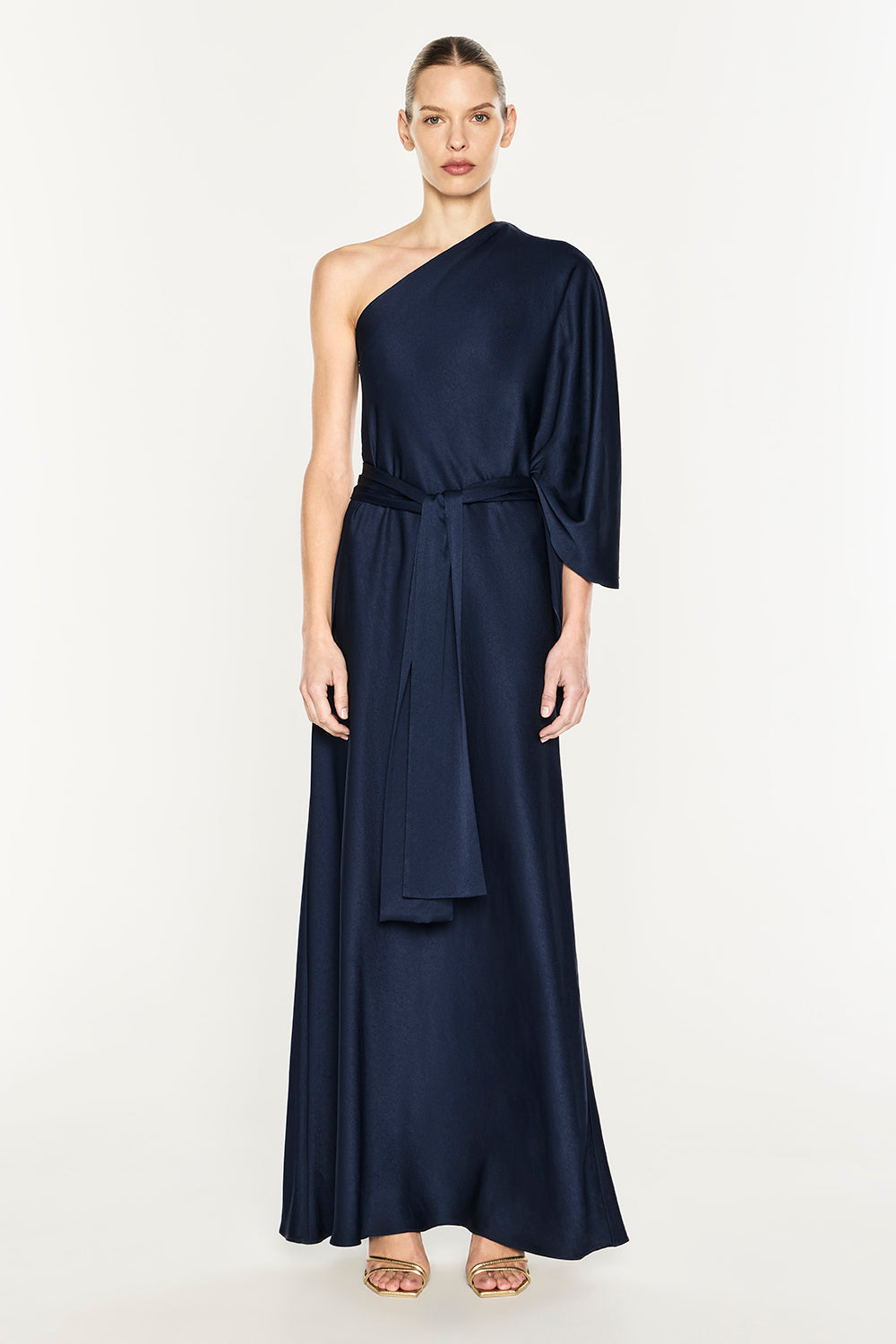 A woman modeling a navy blue one-shoulder maxi dress with a draped sleeve, matching fabric tie belt at the waist, and a flowing skirt, set against a plain light background.