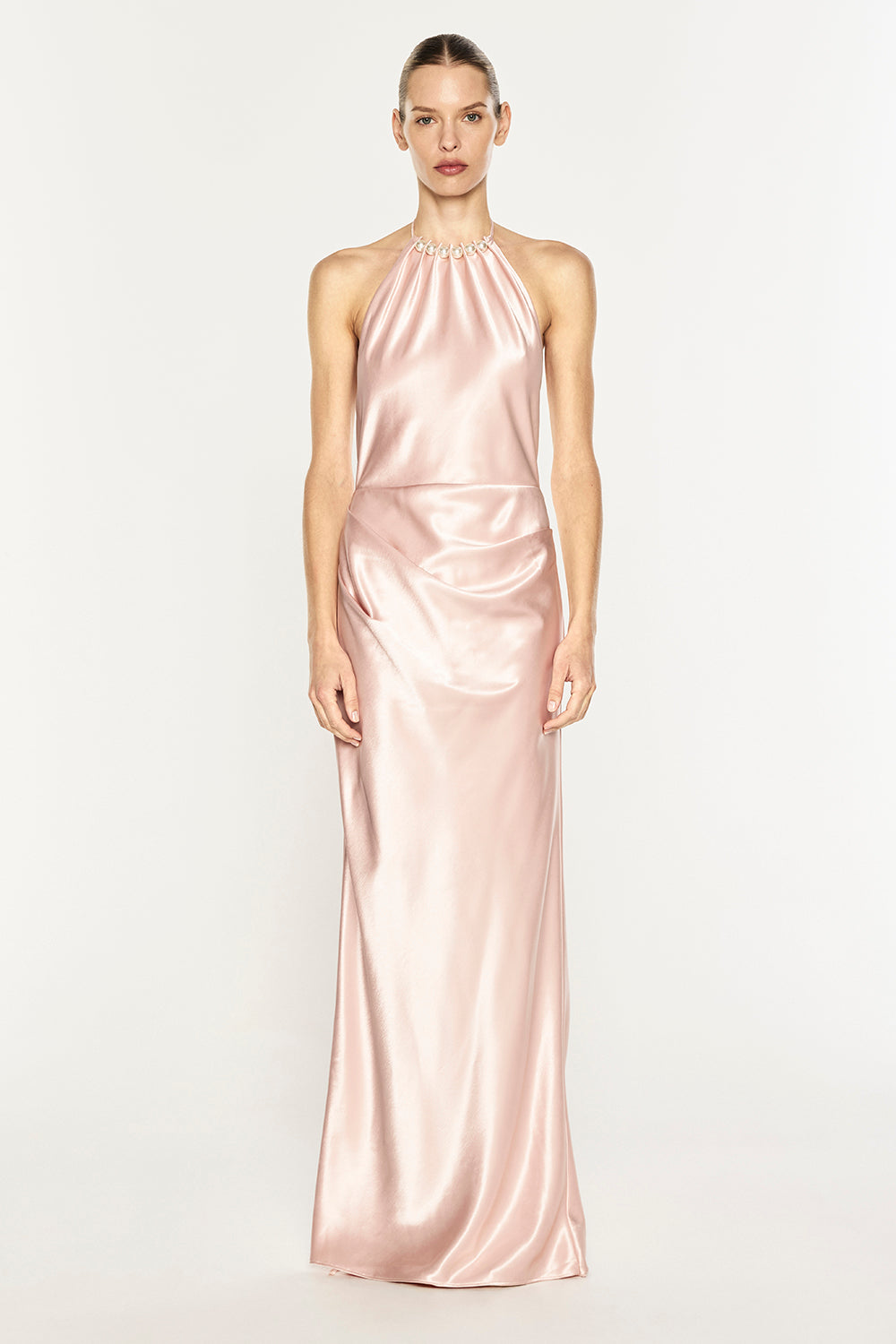 A model wearing a floor-length, satin halter gown in a soft blush pink color, standing against a plain light gray studio background.