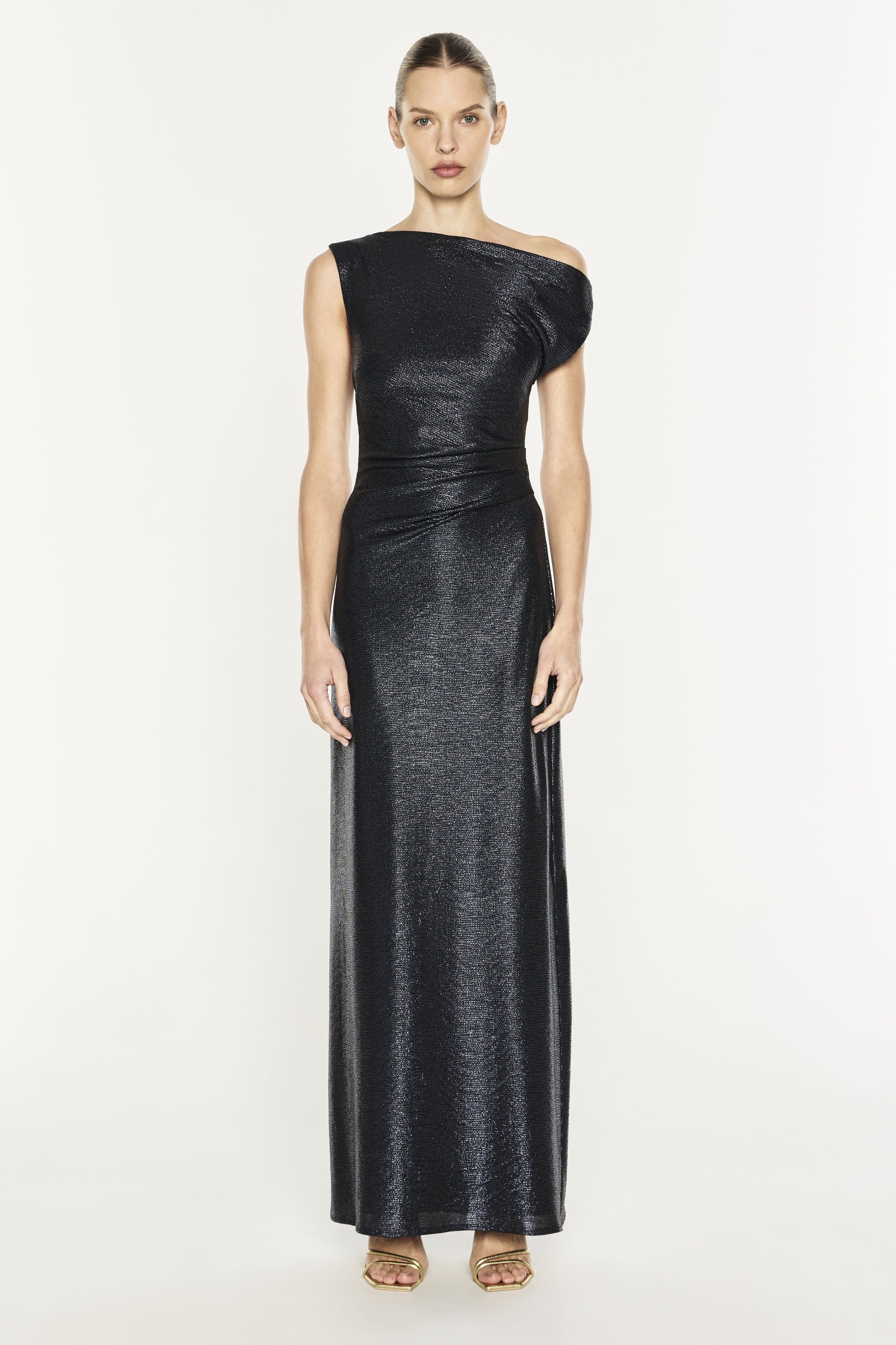 A model wearing a floor-length, one-shoulder black dress with a textured, metallic shimmer, photographed against a solid off-white studio background.