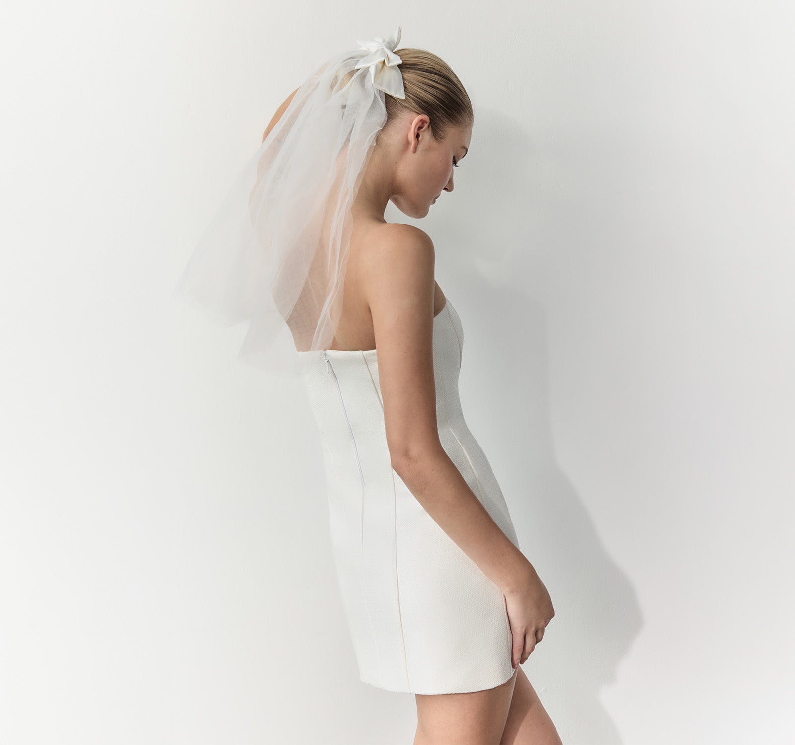 A woman in a strapless white wedding dress with a sheer white veil and a white floral hair accessory, photographed against a plain white wall.
