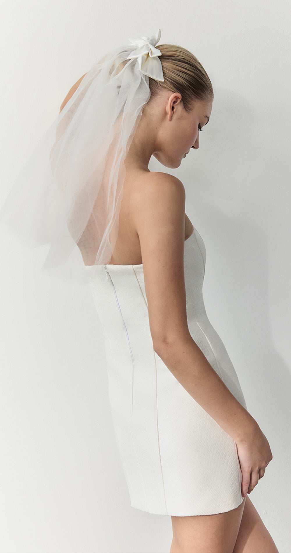 A woman shown from the back wearing a strapless white dress and a sheer white veil with a bow, photographed against a plain light background.