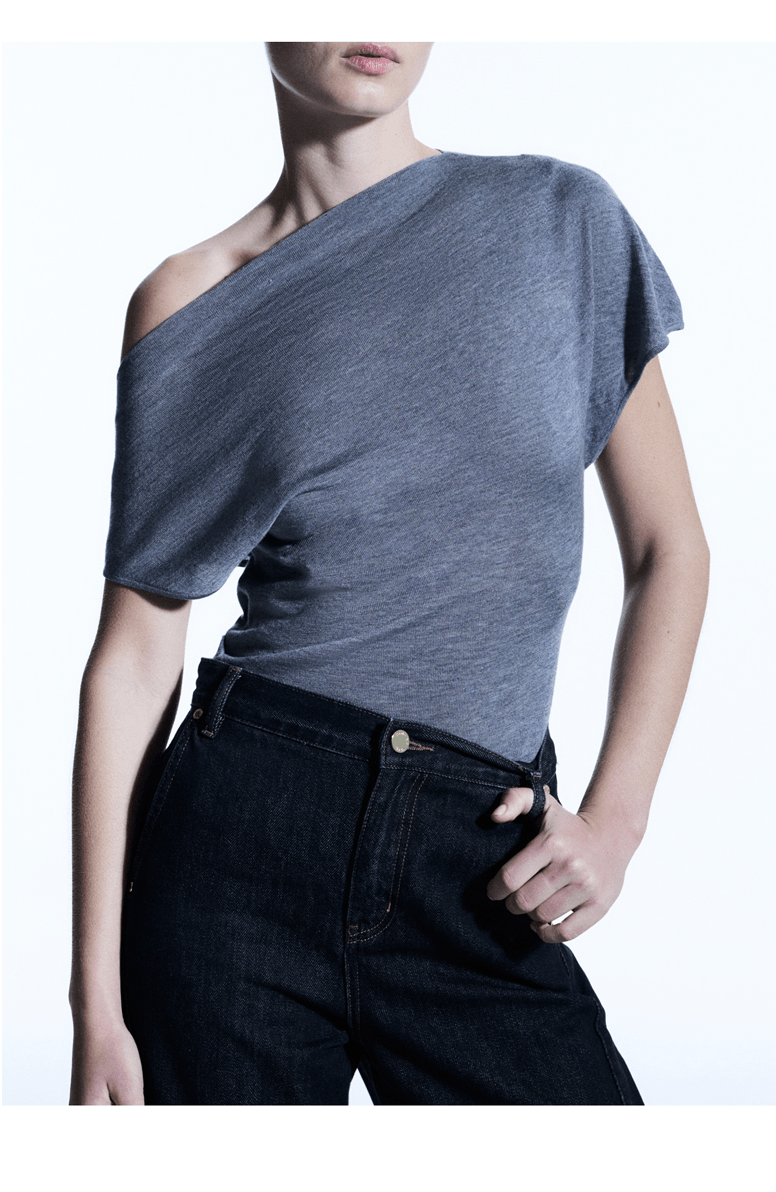 A model wearing a slate blue-gray one-shoulder short-sleeve top and dark jeans, cropped from the lower face to the upper thighs against a plain white studio background.